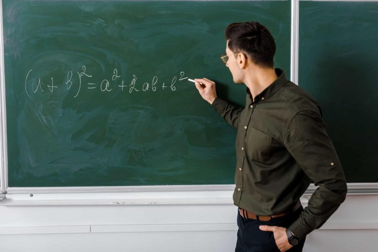 male-teacher-in-formal-wear-writing-equation-on-chalkboard-in-maths-class